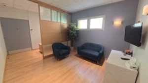 Salon funéraire avec fauteuil et canapé bleus, télévision murale et séparation en bois pour le recueillement.